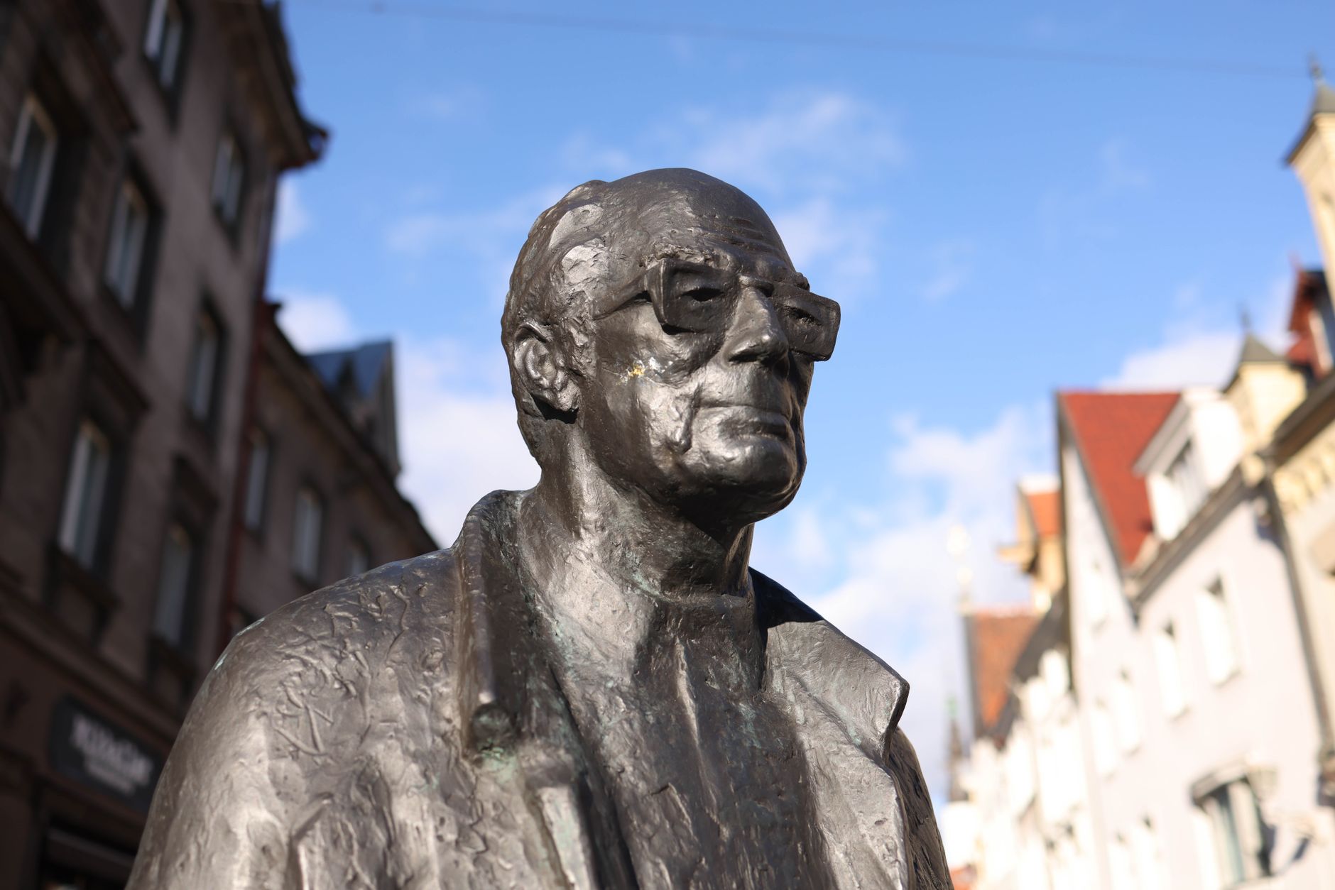 Close-up of a bronze statue wearing eyeglasses, set in a European urban environment with historical architecture.